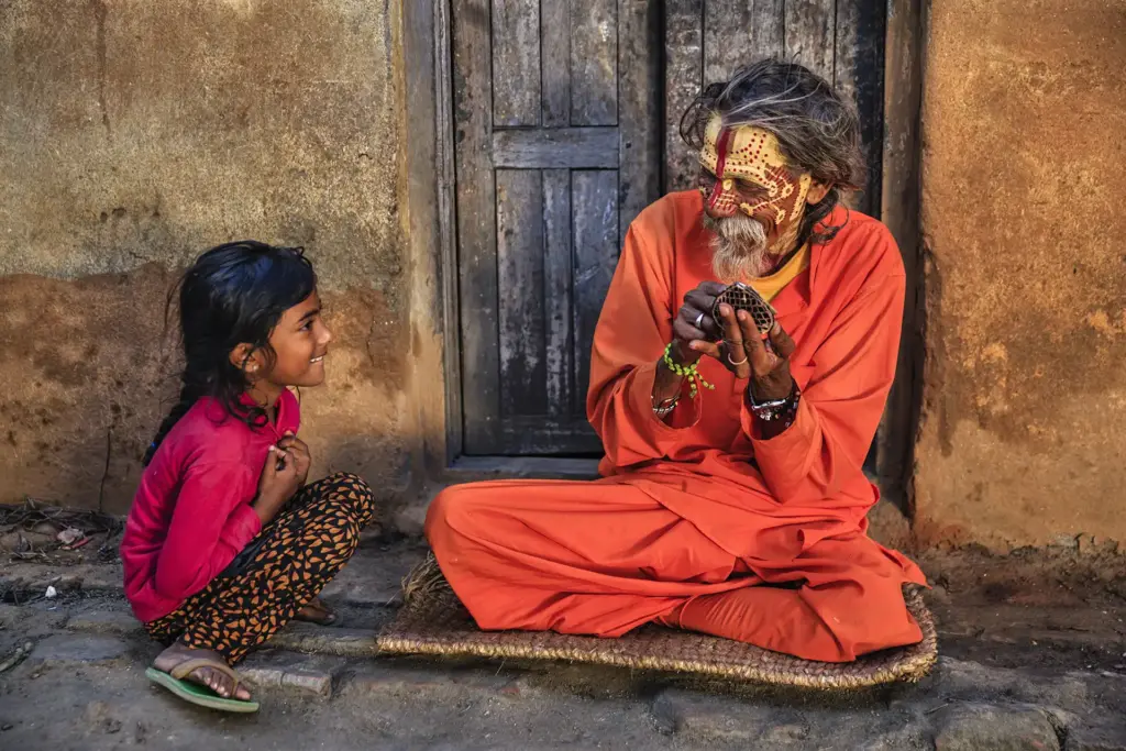 Straßenszene in Indien, ein Guru sitzt im Schneidersitz vor einer alten Haustür, ein indisches Mädchen hockt links von ihm und tritt mit ihm in Interaktion